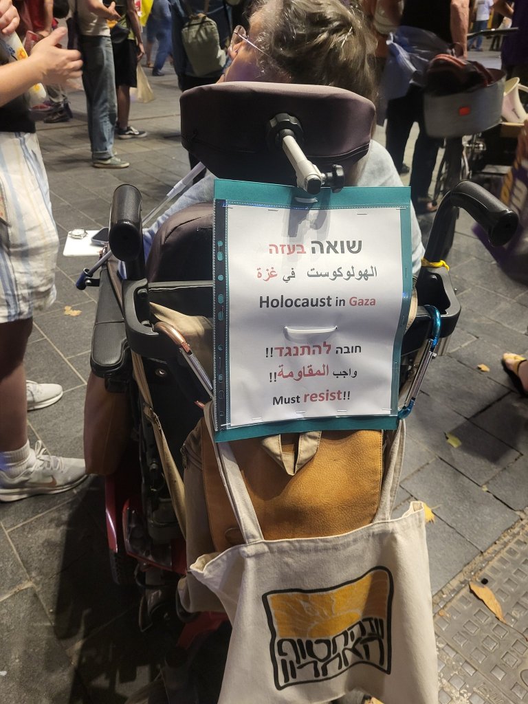 

**Alt text:**
*A person in a wheelchair at a street protest holds a sign on the back of their chair. The sign, written in Hebrew, Arabic, and English, reads: “Holocaust in Gaza. חובה להתנגד!! واجب المقاومة!! Must resist!!” A canvas bag with the logo of the Israeli group “וועד ארגון ההורים” hangs from the wheelchair. Other protesters are visible in the background.*

